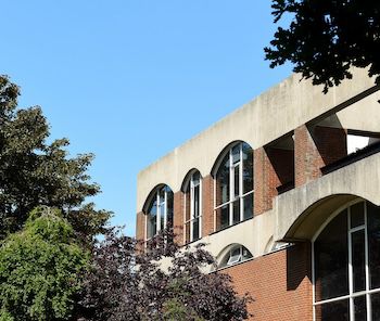 A photograph of a corner of Falmer House at the University of Sussex with trees in the foreground and blue sky above