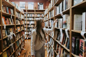 A young woman, with long brown hair and glasses browsing books on wooden shelves with rows of books lining the aisles.
