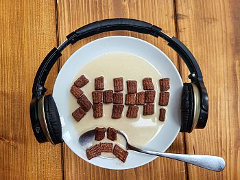 A bowl of cereal with a pair of headphones placed around the edge of the bowl. The cereal in the bowl spells out the word SHH! with a sad face on the spoon.