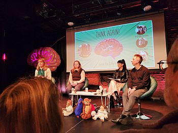 A panel of presenters, including Dr Louisa Rinaldi. A presentation behind them has an image of a brain and text 'Think Again'. There is a display of soft toys.